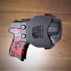 Puede incluir: Una pistola negra y roja con un patr&oacute;n de camuflaje est&aacute; en una funda negra con un patr&oacute;n de fibra de carbono. La funda tiene el texto "Made in the USA" y un dise&ntilde;o de bandera estadounidense.