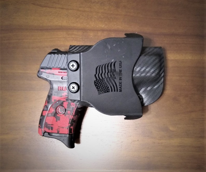 Puede incluir: Una pistola negra y roja con un patr&oacute;n de camuflaje est&aacute; en una funda negra con un patr&oacute;n de fibra de carbono. La funda tiene el texto "Made in the USA" y un dise&ntilde;o de bandera estadounidense.