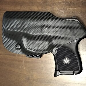 May include: A black handgun with a carbon fiber holster. The gun has a black grip with a silver logo on the side.