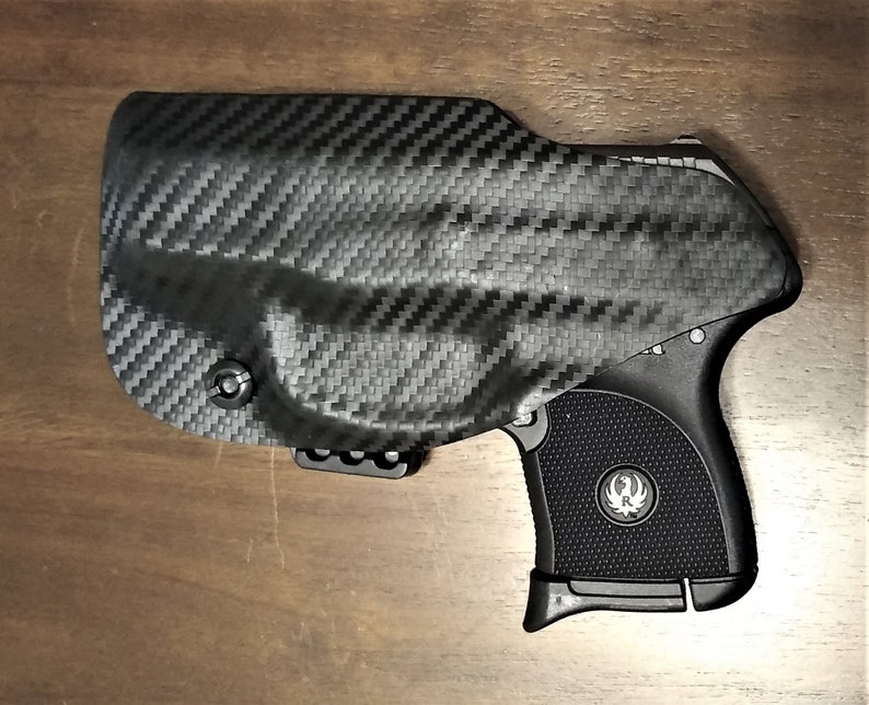 May include: A black handgun with a carbon fiber holster. The gun has a black grip with a silver logo on the side.