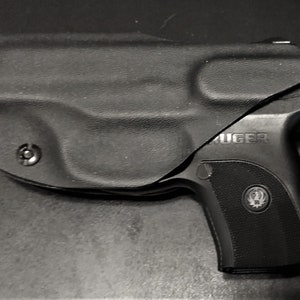 Puede incluir: Una pistola negra con una funda negra. La pistola tiene un logotipo Ruger plateado en el lateral.