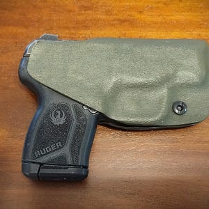 Pu&ograve; includere: Una pistola nera con una fondina verde. La pistola ha un'impugnatura testurizzata e la scritta "RUGER" stampata sul lato.