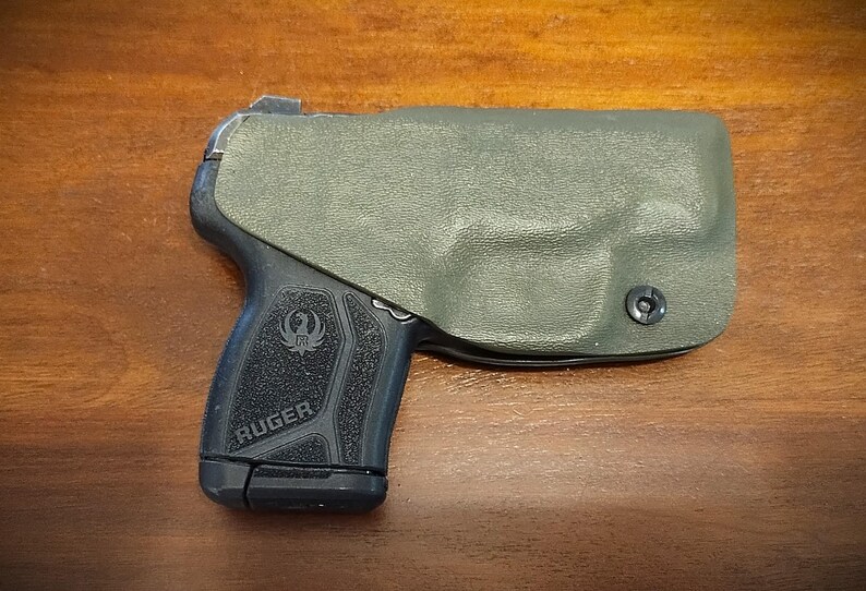 Pu&ograve; includere: Una pistola nera con una fondina verde. La pistola ha un'impugnatura testurizzata e la scritta "RUGER" stampata sul lato.