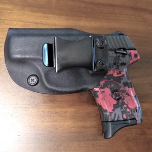 Puede incluir: Una pistola negra con un patr&oacute;n de camuflaje rosa y gris est&aacute; enfundada en una funda de cuero negra con un clip para cintur&oacute;n.