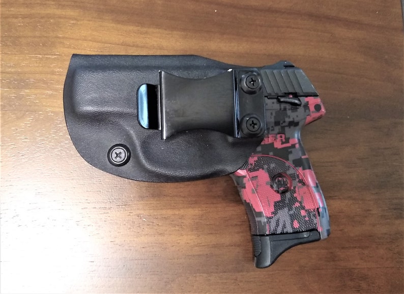 Puede incluir: Una pistola negra con un patr&oacute;n de camuflaje rosa y gris est&aacute; enfundada en una funda de cuero negra con un clip para cintur&oacute;n.