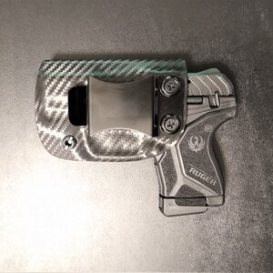 Puede incluir: Una funda de pistola negra y gris con un patr&oacute;n de fibra de carbono y un clip negro. La funda est&aacute; dise&ntilde;ada para adaptarse a una pistola Ruger.