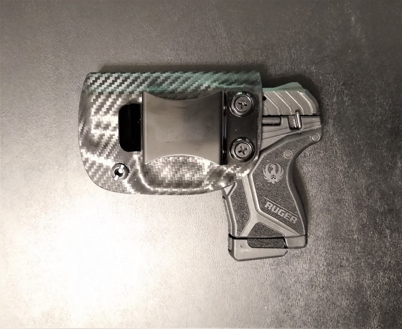 Puede incluir: Una funda de pistola negra y gris con un patr&oacute;n de fibra de carbono y un clip negro. La funda est&aacute; dise&ntilde;ada para adaptarse a una pistola Ruger.