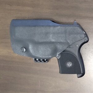 May include: A black handgun with a black holster. The holster has a clip on the bottom and a small button on the side. The gun has a textured grip and a silver logo on the side.
