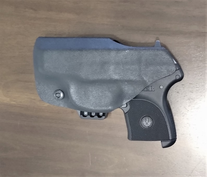 May include: A black handgun with a black holster. The holster has a clip on the bottom and a small button on the side. The gun has a textured grip and a silver logo on the side.