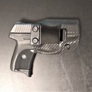 Puede incluir: Una pistola negra y plateada con una funda de fibra de carbono negra. La pistola tiene una empu&ntilde;adura negra y una corredera plateada. La funda tiene un clip para cintur&oacute;n y un patr&oacute;n de fibra de carbono.
