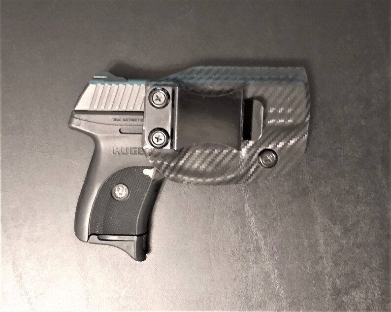 Puede incluir: Una pistola negra y plateada con una funda de fibra de carbono negra. La pistola tiene una empu&ntilde;adura negra y una corredera plateada. La funda tiene un clip para cintur&oacute;n y un patr&oacute;n de fibra de carbono.