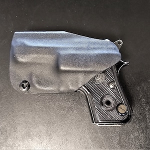 May include: A black handgun with a brown grip is holstered in a black leather holster.