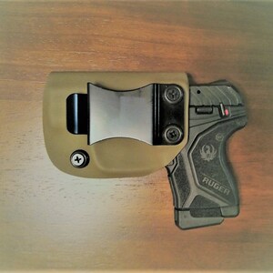 May include: A tan leather holster with a black metal clip and a black Ruger pistol with a textured grip.