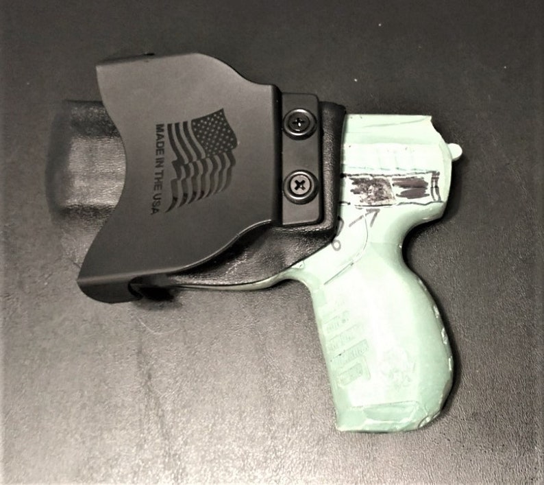 May include: A black leather holster with a  'MADE IN THE USA' inscription and an American flag design. The holster is holding a light green handgun.