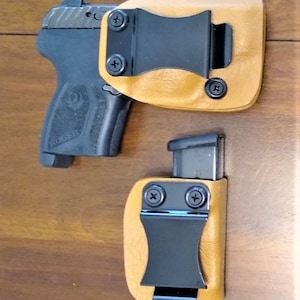 K&ouml;nnte beinhalten: Zwei Lederholster mit schwarzen Metallclips f&uuml;r eine Pistole und ein Magazin. Die Holster sind tanfarben und haben eine strukturierte Oberfl&auml;che.