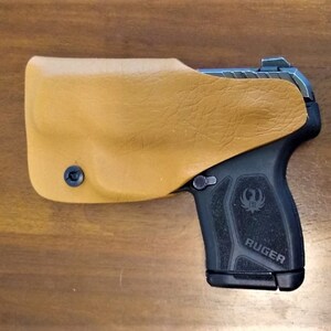 K&ouml;nnte beinhalten: Eine schwarze Ruger-Pistole mit einem braunen Lederholster. Die Pistole hat einen schwarzen Griff mit dem aufgedruckten Wort "RUGER".