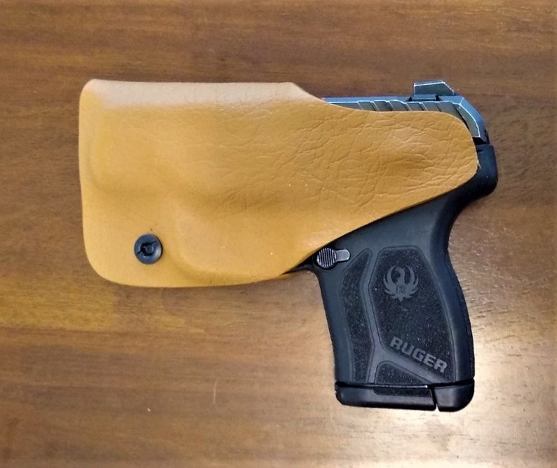 K&ouml;nnte beinhalten: Eine schwarze Ruger-Pistole mit einem braunen Lederholster. Die Pistole hat einen schwarzen Griff mit dem aufgedruckten Wort "RUGER".