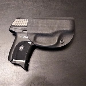 Puede incluir: Una pistola negra con una funda negra. La pistola tiene un deslizador plateado y el texto "RUGER" es visible en el lateral.