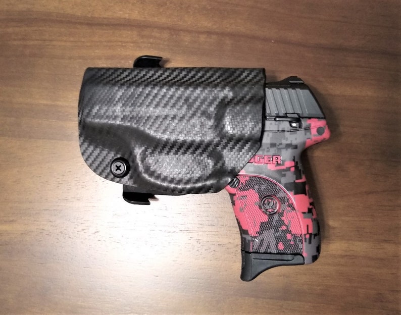 Puede incluir: Una pistola con estampado negro y rojo con una funda de fibra de carbono negra. La funda tiene un tornillo en el lateral.