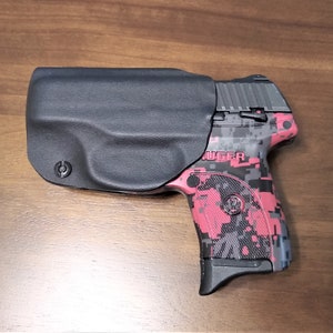 Puede incluir: Una funda de cuero negra con una pistola Ruger con estampado de camuflaje rojo y negro. La funda tiene un logotipo circular en el lateral.