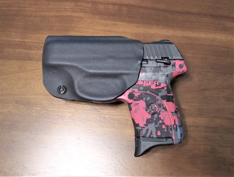 Puede incluir: Una funda de cuero negra con una pistola Ruger con estampado de camuflaje rojo y negro. La funda tiene un logotipo circular en el lateral.