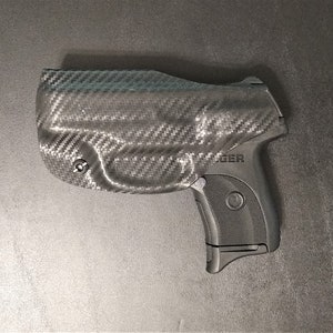 Puede incluir: Una pistola negra y gris con una funda de fibra de carbono. La funda tiene un peque&ntilde;o tornillo en el lateral. La pistola tiene la palabra "ROGER" impresa en el lateral.