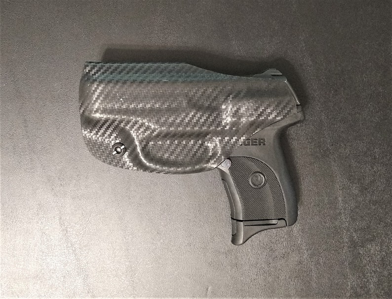 Puede incluir: Una pistola negra y gris con una funda de fibra de carbono. La funda tiene un peque&ntilde;o tornillo en el lateral. La pistola tiene la palabra "ROGER" impresa en el lateral.