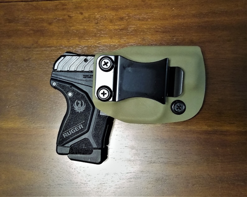 May include: A black and green handgun with a black holster and a clip. The holster is made of a green material and has a black clip. The handgun has a black grip and a silver slide. The handgun is a Ruger.