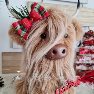 Puede incluir: Un peluche de vaca de las Tierras Altas con largo pelaje marr&oacute;n, adornado con un lazo escoc&eacute;s rojo y verde y una etiqueta de madera "Christmas". La vaca tiene ojos negros y cuernos curvados, para la decoraci&oacute;n navide&ntilde;a.