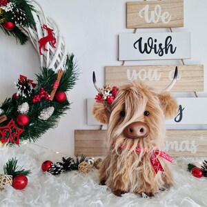 Puede incluir: Un peluche de vaca de las Tierras Altas con largo y lanudo pelaje marr&oacute;n, con una cinta roja. Una corona navide&ntilde;a y un letrero de madera que dice "We Wish You a Merry Christmas" est&aacute;n en el fondo. La vaca est&aacute; sentada sobre una superficie blanca y esponjosa.