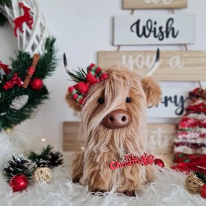 Puede incluir: Un peluche de vaca de las Tierras Altas marr&oacute;n con pelo largo y lanudo, con un lazo escoc&eacute;s rojo y verde. Tiene una nariz marr&oacute;n y peque&ntilde;os cuernos. Un adorno navide&ntilde;o rojo est&aacute; unido a la vaca. El fondo incluye un &aacute;rbol de Navidad y una corona.