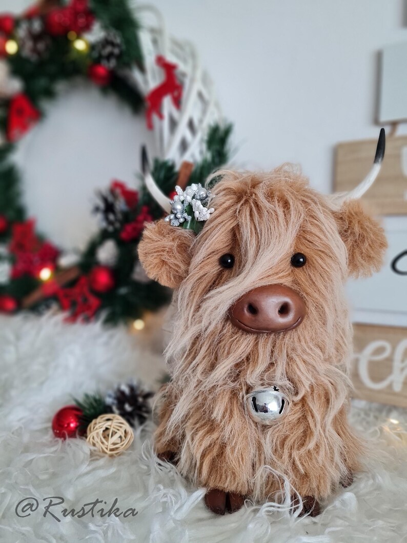 Puede incluir: Un peluche de vaca de las Tierras Altas marr&oacute;n con pelo largo y lanudo, una campana plateada y peque&ntilde;os cuernos blancos. La vaca tiene una nariz marr&oacute;n y est&aacute; decorada con un peque&ntilde;o adorno floral. El fondo incluye una corona y adornos navide&ntilde;os.