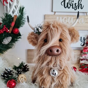 Puede incluir: Un peluche de vaca de las Tierras Altas con pelo largo y lanudo de color marr&oacute;n, una campana plateada y peque&ntilde;os cuernos. La vaca est&aacute; decorada con una peque&ntilde;a corona floral y se sienta sobre una superficie blanca y esponjosa. Decoraciones navide&ntilde;as en el fondo.