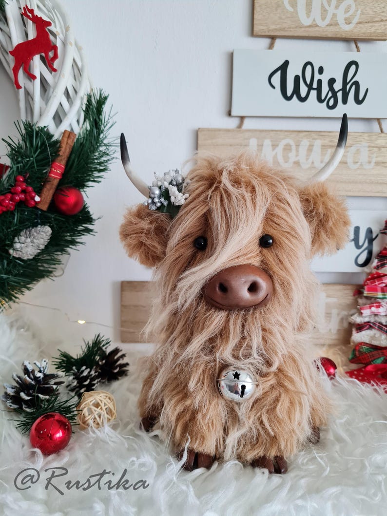 Puede incluir: Un peluche de vaca de las Tierras Altas con pelo largo y lanudo de color marr&oacute;n, una campana plateada y peque&ntilde;os cuernos. La vaca est&aacute; decorada con una peque&ntilde;a corona floral y se sienta sobre una superficie blanca y esponjosa. Decoraciones navide&ntilde;as en el fondo.