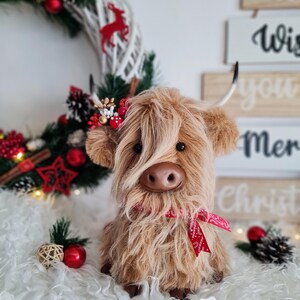 Puede incluir: Un peluche de vaca de las Tierras Altas con pelo largo y lanudo de color marr&oacute;n, una cinta roja y peque&ntilde;os cuernos. La vaca est&aacute; colocada frente a una corona navide&ntilde;a y elementos decorativos, incluyendo adornos rojos y un letrero que dice "Wish you Merry Christmas".