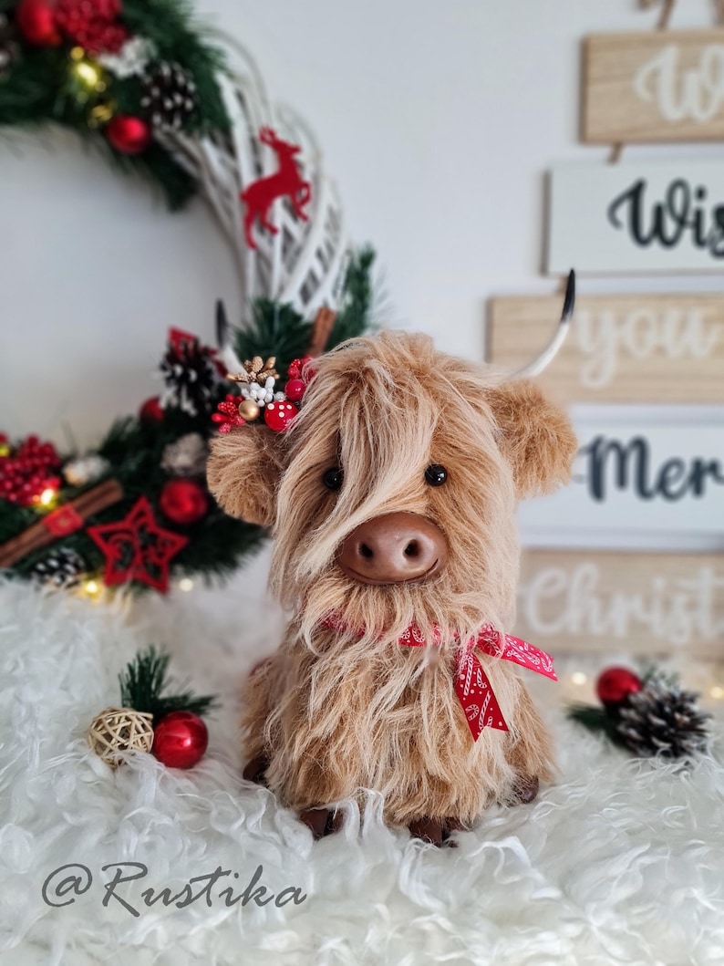 Puede incluir: Un peluche de vaca de las Tierras Altas con pelo largo y lanudo de color marr&oacute;n, una cinta roja y peque&ntilde;os cuernos. La vaca est&aacute; colocada frente a una corona navide&ntilde;a y elementos decorativos, incluyendo adornos rojos y un letrero que dice "Wish you Merry Christmas".