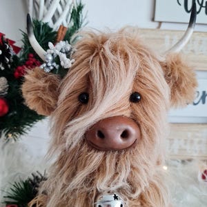 Puede incluir: Una vaca de las Tierras Altas de peluche con pelo largo y lanudo de color marr&oacute;n claro, ojos negros y una nariz marr&oacute;n. Tiene cuernos peque&ntilde;os y curvados y una campana plateada alrededor del cuello. La vaca est&aacute; decorada con una corona navide&ntilde;a y adornos.
