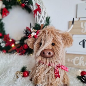Puede incluir: Una vaca de las Tierras Altas de peluche con pelo largo y lanudo de color marr&oacute;n, una cinta roja y una diadema floral. La vaca est&aacute; sentada sobre una superficie blanca y esponjosa, con una corona navide&ntilde;a y decoraciones en el fondo. El texto "@Rustika" es visible.