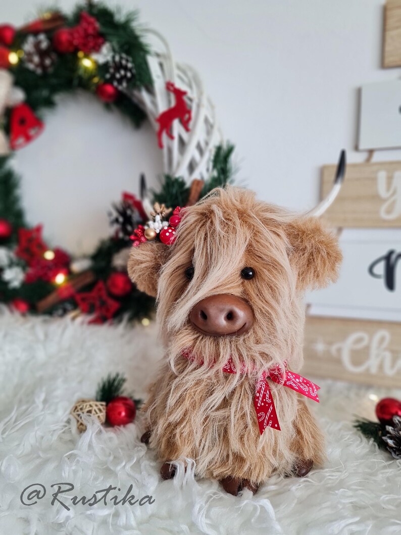Puede incluir: Una vaca de las Tierras Altas de peluche con pelo largo y lanudo de color marr&oacute;n, una cinta roja y una diadema floral. La vaca est&aacute; sentada sobre una superficie blanca y esponjosa, con una corona navide&ntilde;a y decoraciones en el fondo. El texto "@Rustika" es visible.