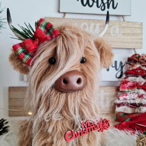 Puede incluir: Un peluche de vaca de las Tierras Altas con largo y desgre&ntilde;ado pelaje marr&oacute;n, adornado con un lazo escoc&eacute;s rojo y verde. Tiene peque&ntilde;os ojos negros, una nariz marr&oacute;n y peque&ntilde;os cuernos. Un adorno navide&ntilde;o rojo cuelga de su cuello. El fondo incluye letreros de madera con la palabra "Wish".