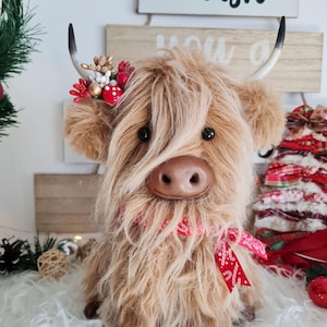 Puede incluir: Un peluche de vaca de las Tierras Altas con largo y lanudo pelaje marr&oacute;n, ojos negros y nariz marr&oacute;n. Lleva una bufanda con estampado rojo y blanco y un adorno festivo para la cabeza con bayas rojas y detalles dorados. Se muestra la palabra "Wish".