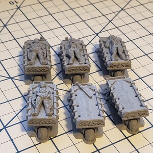 May include: Six gray miniature figures are lying on wheeled platforms. The figures are in various poses and appear to be bound. The platforms are detailed with carvings and have wheels.
