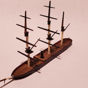 May include: A wooden model of a tall ship with three masts and multiple sails. The ship has a brown hull and deck, with black masts and rigging. The model is set against a white background.