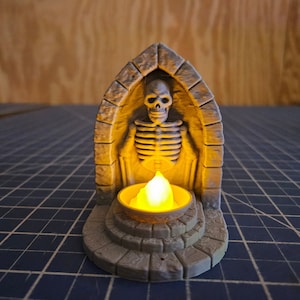 Puede incluir: Una pequeña estructura de piedra gris con una figura esquelética en su interior. La estructura tiene una puerta arqueada y un pequeño portavelas en frente. La vela está encendida y brilla en amarillo.