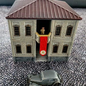 May include: A gray, two-story building with a red roof and a red flag with a star hanging from the balcony. A small, gray car is parked in front of the building.