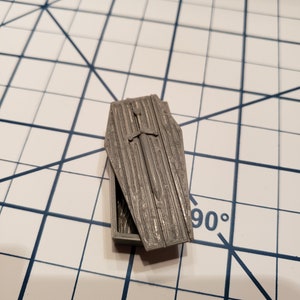 May include: A small, gray, 3D printed coffin with a sword inside. The coffin is open and the sword is visible.