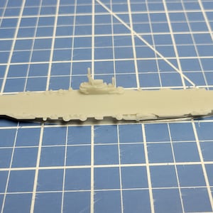 May include: A light gray plastic model of an aircraft carrier. The model has a flat deck with a small tower and several details along the sides. The model is placed on a blue grid surface.