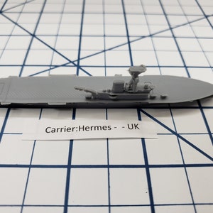 May include: A grey plastic model of the HMS Hermes aircraft carrier. The model is detailed with a flight deck, a small island, and a radar mast. The text "Carrier: Hermes - UK" is printed on a white label below the model.