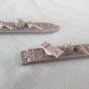 Puede incluir: Dos modelos de barcos de plástico gris, uno con un lanzamisiles elevado, el otro con una torreta de cañón. Los barcos están sobre una superficie gris claro.