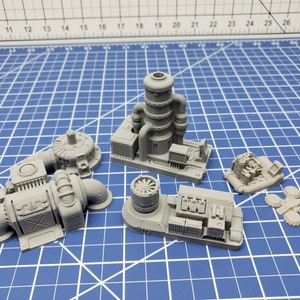 May include: Five gray 3D printed miniature models of industrial structures. The models are made of a smooth, gray plastic and feature intricate details, including pipes, gears, and other industrial elements. The models are arranged on a blue grid surface.